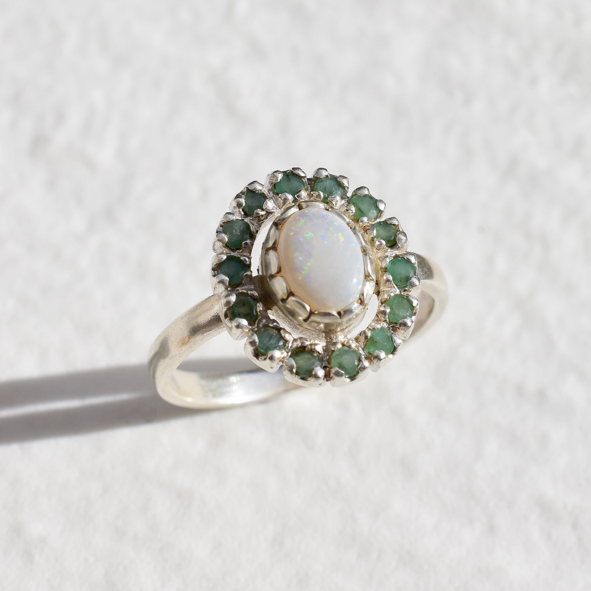 Australian Opal and Colombian Emerald halo ring in sterling silver with oval cabochon opal and diamond-cut emeralds, perfect for statement elegance, believed to inspire love and prosperity, an ideal October and May birthstone gift for weddings, anniversaries, and special celebrations.