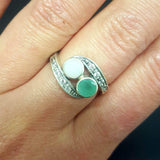 Bypass style ring featuring a natural Green Emerald and a cabochon White Opal in solid 925 sterling silver, accented with CZ pave stones for added brilliance. The combination of real Emerald and Opal highlights vibrant green tones and flashes of fire colors. With Emerald as the May Birthstone and Opal as the October Birthstone, this two stone design makes a meaningful gift for birthdays, anniversaries, and personal milestones. Perfect to wear at weddings, evening parties, and special celebrations.