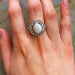 Opal Bezel Halo Ring worn on Middle  finger, showcasing an oval opal with rainbow and fire colors surrounded by CZ diamonds. Perfect for special occasions, weddings and cocktail parties. Ideal gift for October babies.