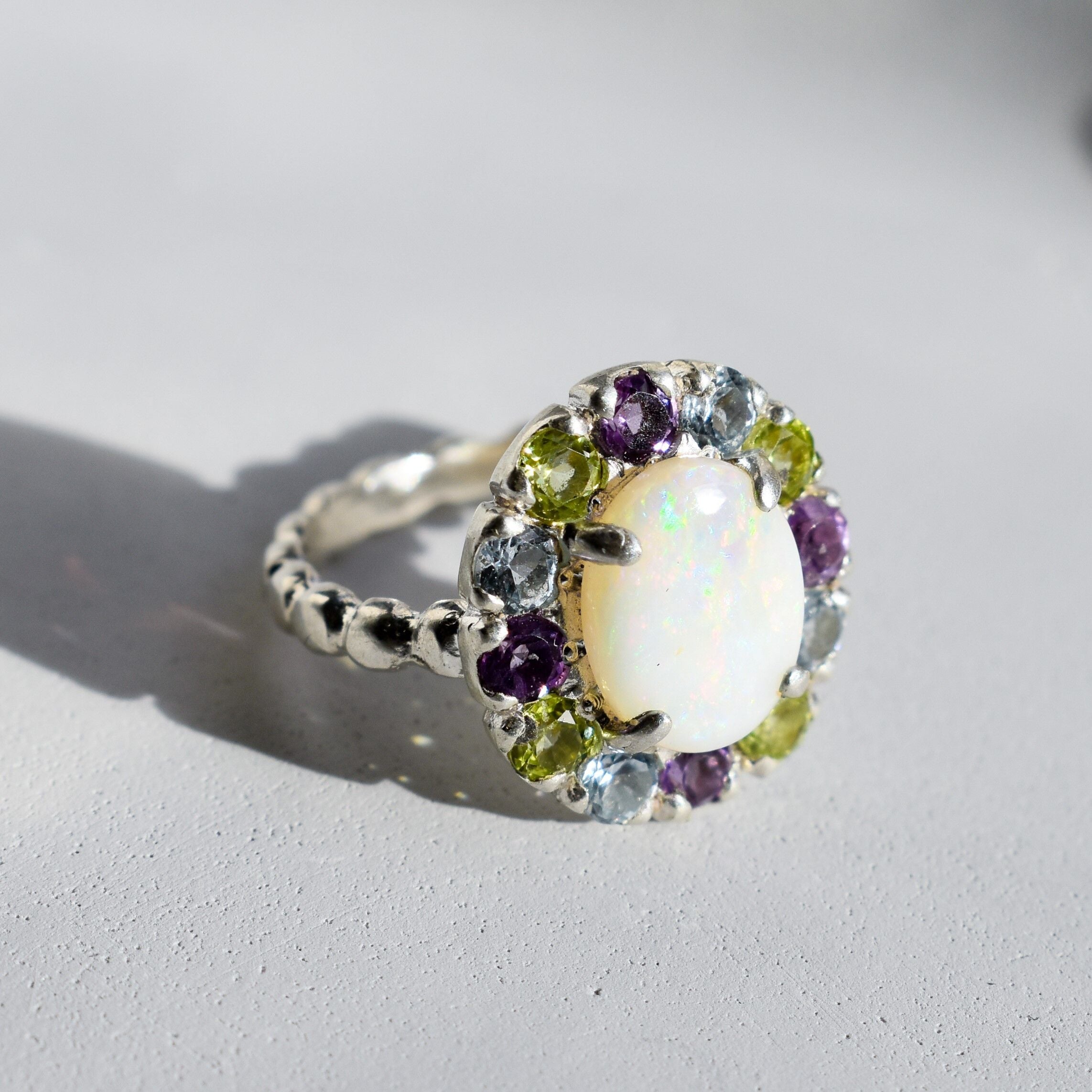 White Opal flower cluster ring in sterling silver with colorful Amethyst and Peridot halo in a cocktail style. The elegant cluster design adds sparkle. Opal is linked with hope and clarity, Amethyst with calm, Peridot with fresh energy. A lovely birthstone gift for October, February and August. Wear to weddings, graduations, dinner dates and holiday parties.