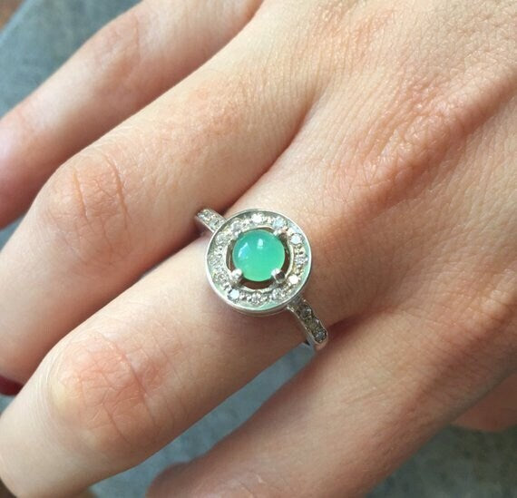 Emerald Halo Ring worn on the index finger with glossy red nails, sterling silver band accented with CZ stones highlighting the central opal’s flashes of fire colors. A chic October Birthstone design perfect for evening parties, romantic dinners, and wedding receptions.