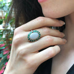 Flower Opal Ring with emerald halo worn on index finger with nude nails, a bold cocktail statement piece that enhances confidence, meaningful as an October and May birthstone gift, perfect for weddings and gala events.