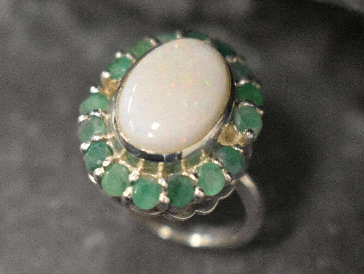 Opal and Emerald Large Cocktail Ring in sterling silver featuring an oval white opal cabochon framed with genuine emerald petals, a statement jewelry style that radiates flashes of fire colors while symbolizing love and renewal, making it a meaningful October and May birthstone gift idea. Perfect for weddings, cocktail parties, and formal evening events where a bold elegant look is desired.