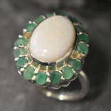Opal and Emerald Large Cocktail Ring in sterling silver featuring an oval white opal cabochon framed with genuine emerald petals, a statement jewelry style that radiates flashes of fire colors while symbolizing love and renewal, making it a meaningful October and May birthstone gift idea. Perfect for weddings, cocktail parties, and formal evening events where a bold elegant look is desired.