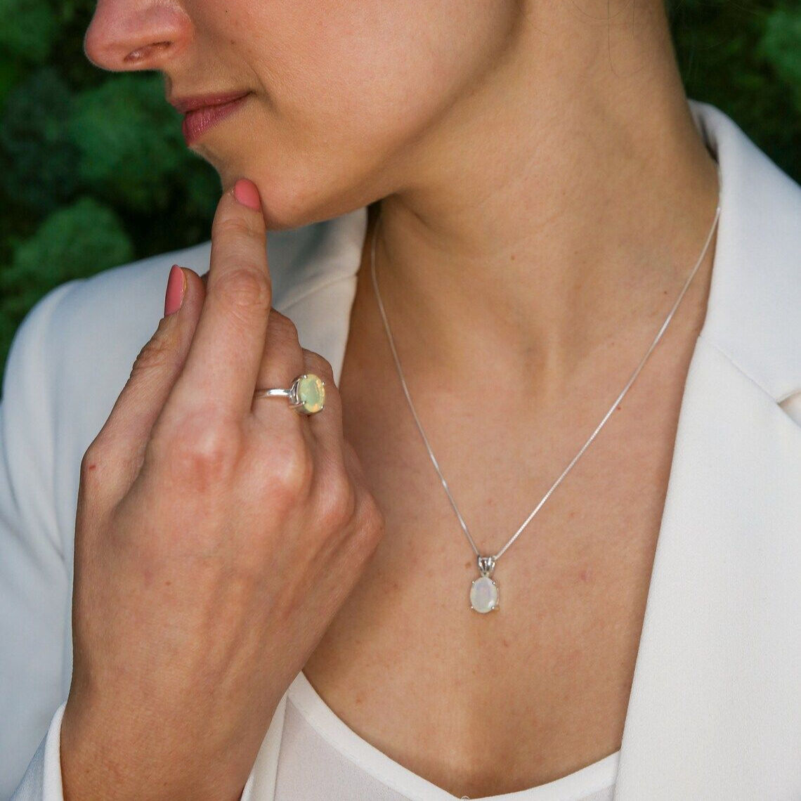 Ethiopian Opal Ring worn on middle finger with pink nails and matching Ethiopian Opal Pendant Necklace styled with a white blazer. The cabochon cut opals show flashes of fire colors set in sterling silver. A classic and elegant jewelry set perfect for October birthstone gifting, anniversaries, promise occasions, and formal events.