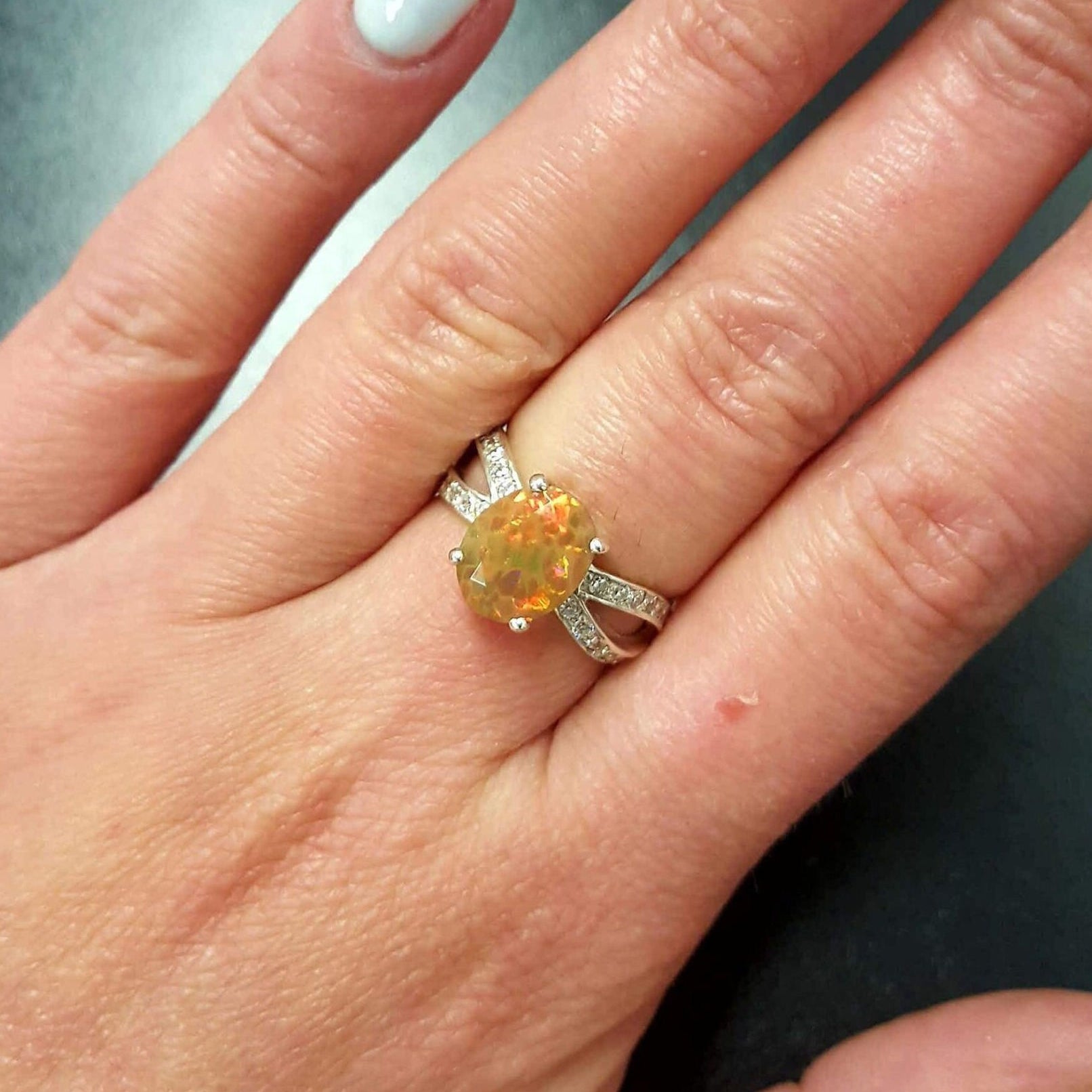 Opal Oval Ring worn on middle finger with natural nails. Jewelry style is bold and refined with a split shank band and 4 prong setting. The gemstone shows flashes of fire colors with added sparkle from the CZ pave band. Brings creativity, inspiration, and balance. A thoughtful birthstone gift for October birthdays and anniversaries. Ideal for weddings, family gatherings, and formal dinners.