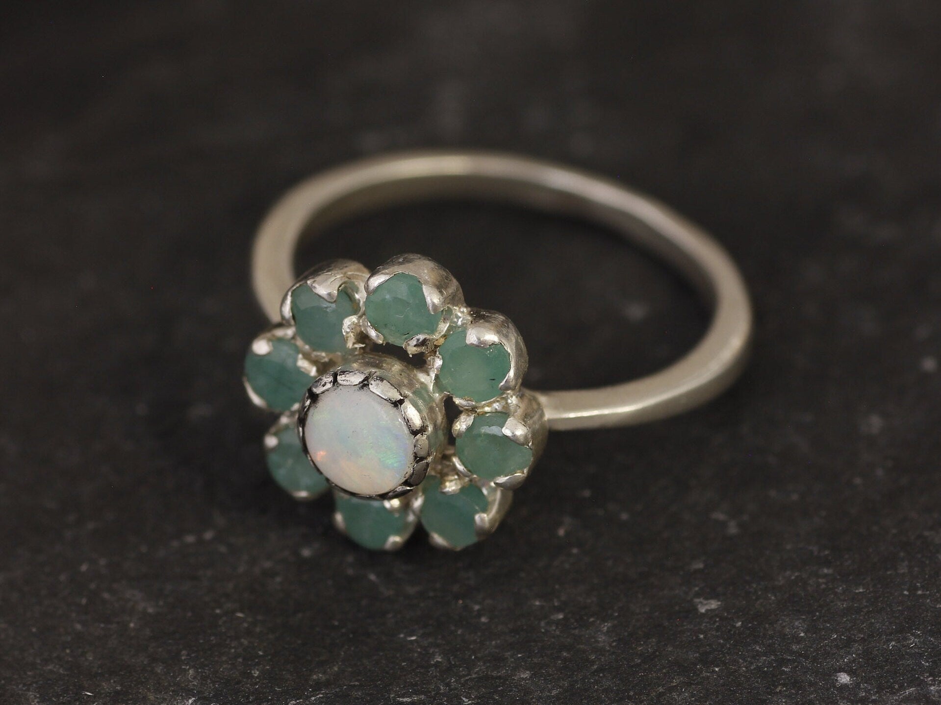 Opal Emerald Flower Ring worn on middle finger with light blue gradient nails. The opal shows flashes of fire colors surrounded by genuine emeralds in a floral cluster design. Perfect for adding inspiration and balance as an October birthstone and symbolizing love and growth as a May birthstone. Suitable for parties, birthdays, or casual outings.