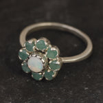 Opal Emerald Flower Ring worn on middle finger with light blue gradient nails. The opal shows flashes of fire colors surrounded by genuine emeralds in a floral cluster design. Perfect for adding inspiration and balance as an October birthstone and symbolizing love and growth as a May birthstone. Suitable for parties, birthdays, or casual outings.
