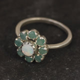 Opal Emerald Flower Ring worn on middle finger with light blue gradient nails. The opal shows flashes of fire colors surrounded by genuine emeralds in a floral cluster design. Perfect for adding inspiration and balance as an October birthstone and symbolizing love and growth as a May birthstone. Suitable for parties, birthdays, or casual outings.