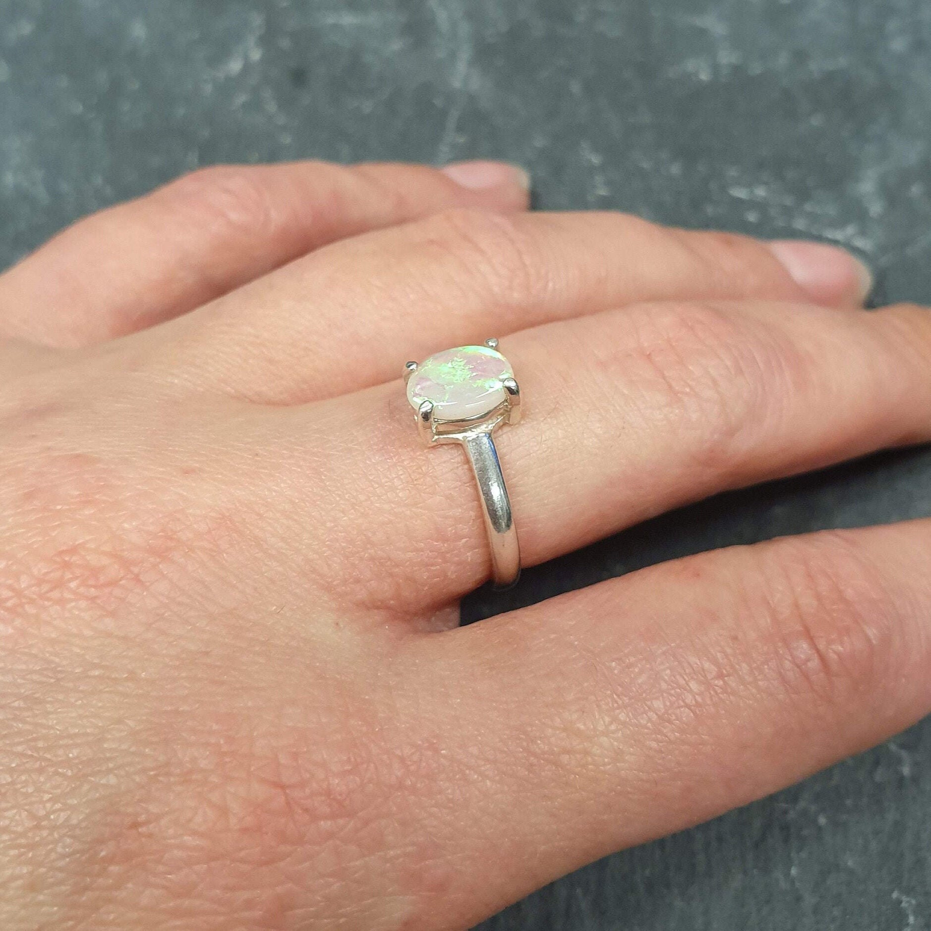 Dainty Opal Engagement Ring styled on middle finger with natural nude nails, ideal for minimalist outfits, believed to inspire creativity and joy, perfect for anniversaries and intimate occasions.
