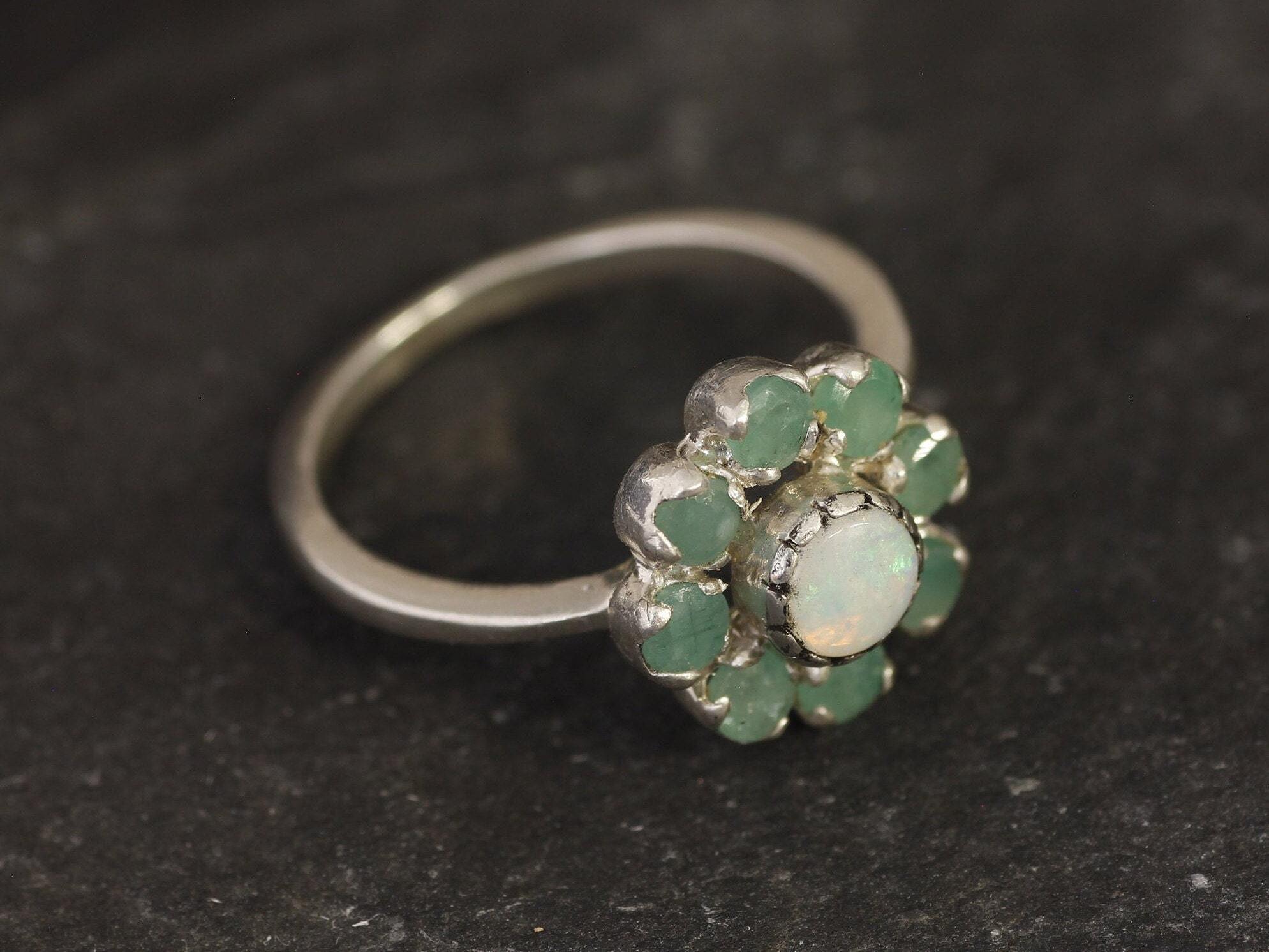Opal Emerald Flower Ring worn on middle finger with light blue gradient nails. The opal shows flashes of fire colors surrounded by genuine emeralds in a floral cluster design. Perfect for adding inspiration and balance as an October birthstone and symbolizing love and growth as a May birthstone. Suitable for parties, birthdays, or casual outings.