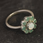 Opal Emerald Flower Ring worn on middle finger with light blue gradient nails. The opal shows flashes of fire colors surrounded by genuine emeralds in a floral cluster design. Perfect for adding inspiration and balance as an October birthstone and symbolizing love and growth as a May birthstone. Suitable for parties, birthdays, or casual outings.