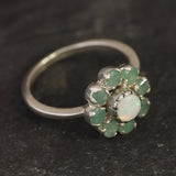 Opal Emerald Flower Ring worn on middle finger with light blue gradient nails. The opal shows flashes of fire colors surrounded by genuine emeralds in a floral cluster design. Perfect for adding inspiration and balance as an October birthstone and symbolizing love and growth as a May birthstone. Suitable for parties, birthdays, or casual outings.