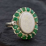 Opal and Emerald Large Cocktail Ring in sterling silver featuring an oval white opal cabochon framed with genuine emerald petals, a statement jewelry style that radiates flashes of fire colors while symbolizing love and renewal, making it a meaningful October and May birthstone gift idea. Perfect for weddings, cocktail parties, and formal evening events where a bold elegant look is desired.