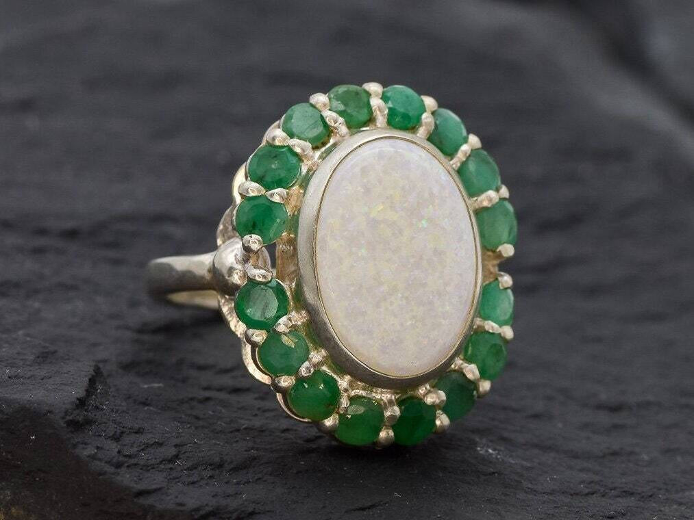 Opal and Emerald Large Cocktail Ring in sterling silver featuring an oval white opal cabochon framed with genuine emerald petals, a statement jewelry style that radiates flashes of fire colors while symbolizing love and renewal, making it a meaningful October and May birthstone gift idea. Perfect for weddings, cocktail parties, and formal evening events where a bold elegant look is desired.
