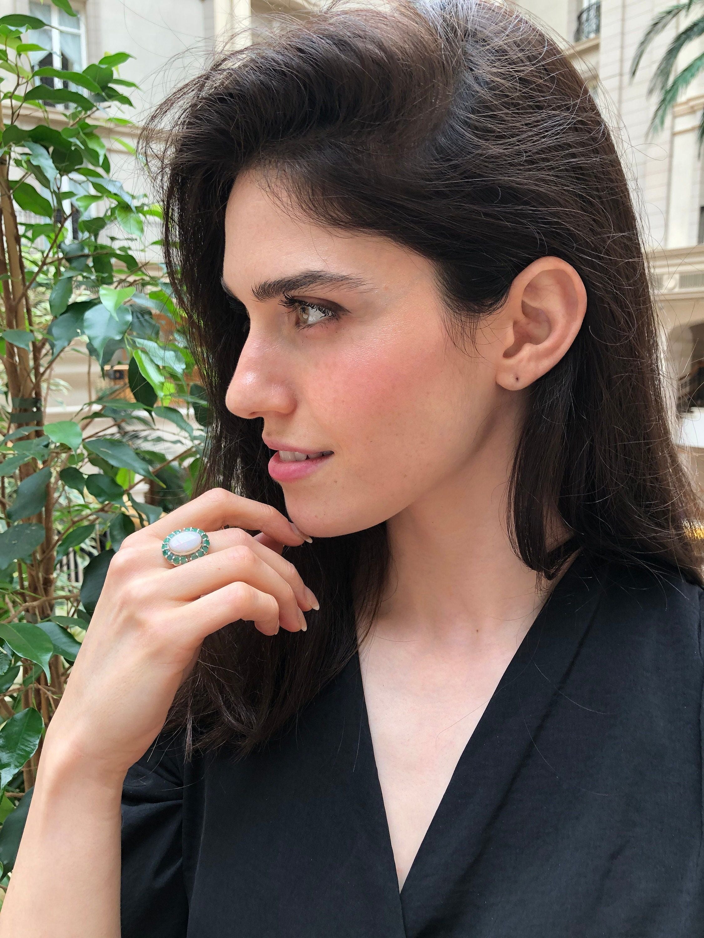 Flower Opal Ring with white opal cabochon styled on middle finger with bare nails, accented by genuine emerald petals for a chic look, symbolic as an October and May birthstone gift, ideal for anniversary dinners and evening celebrations.