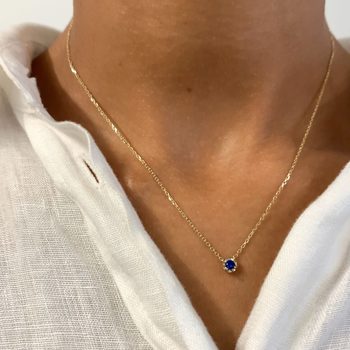 Woman in white linen top, wearing a dainty round sapphire necklace crafted in Solid 14k Yellow Gold chain. Elegant and minimalist blue sapphire september birthstone for everyday wear.
