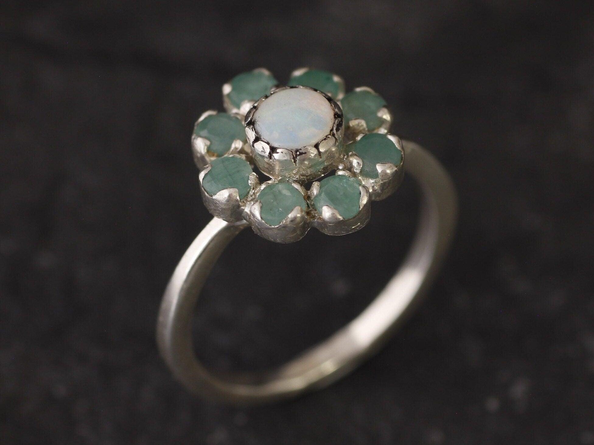 Opal Emerald Flower Ring worn on middle finger with light blue gradient nails. The opal shows flashes of fire colors surrounded by genuine emeralds in a floral cluster design. Perfect for adding inspiration and balance as an October birthstone and symbolizing love and growth as a May birthstone. Suitable for parties, birthdays, or casual outings.
