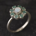 Opal Emerald Flower Ring worn on middle finger with light blue gradient nails. The opal shows flashes of fire colors surrounded by genuine emeralds in a floral cluster design. Perfect for adding inspiration and balance as an October birthstone and symbolizing love and growth as a May birthstone. Suitable for parties, birthdays, or casual outings.
