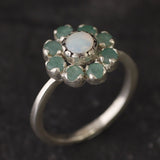 Opal Emerald Flower Ring worn on middle finger with light blue gradient nails. The opal shows flashes of fire colors surrounded by genuine emeralds in a floral cluster design. Perfect for adding inspiration and balance as an October birthstone and symbolizing love and growth as a May birthstone. Suitable for parties, birthdays, or casual outings.