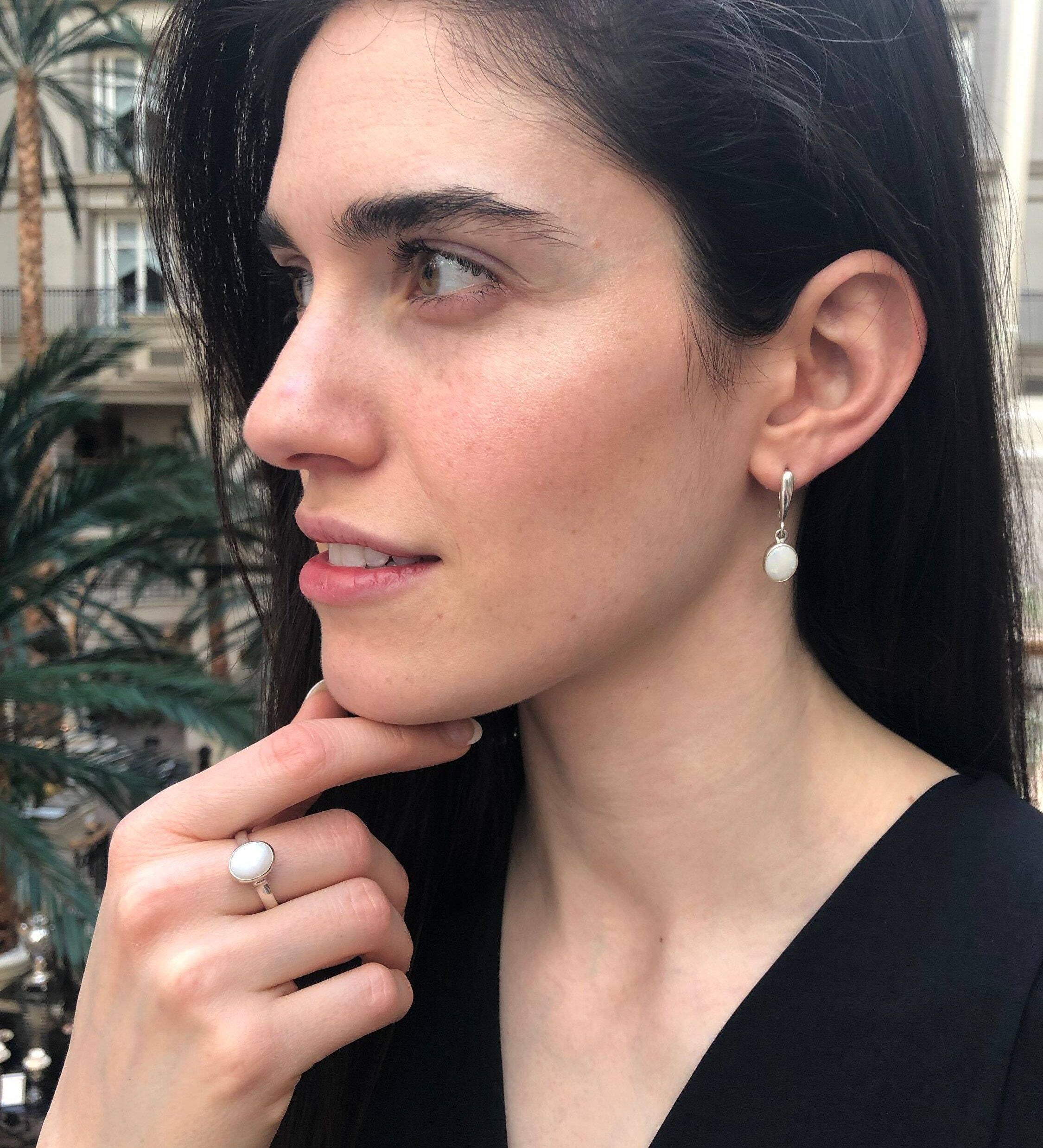 Opal Solitaire Ring styled with a black v-neck blouse and matching white opal drop earrings. Jewelry style is elegant and modern with a bezel design showing flashes of rainbow colors. Symbolizes creativity, harmony, and positivity. A meaningful October birthstone gift for birthdays or milestones. Ideal for evening dinners, weddings, and cocktail events.