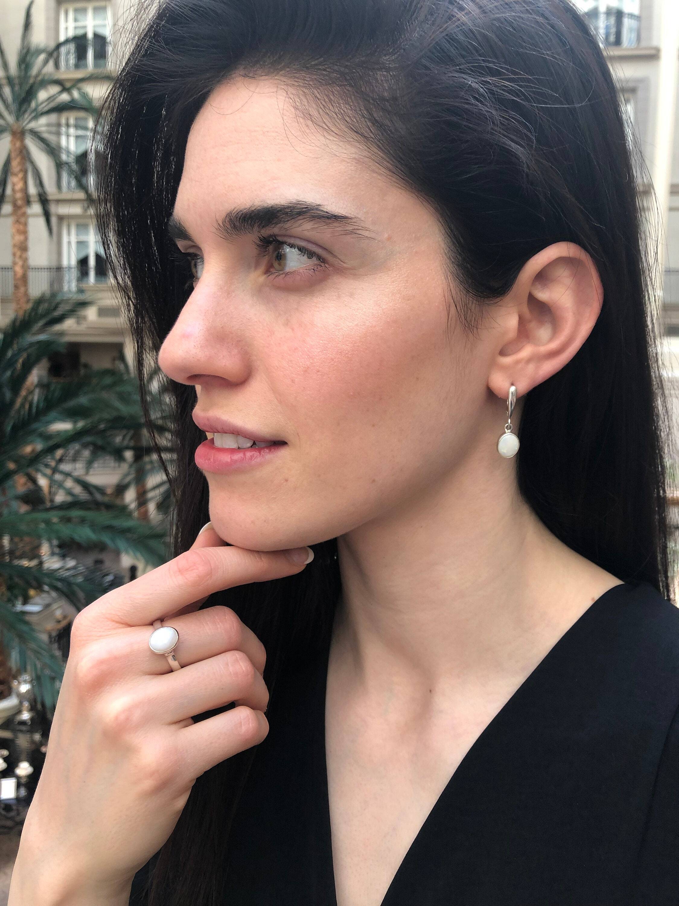 Opal Solitaire Ring styled with a black v-neck blouse and matching white opal drop earrings. Jewelry style is elegant and modern with a bezel design showing flashes of rainbow colors. Symbolizes creativity, harmony, and positivity. A meaningful October birthstone gift for birthdays or milestones. Ideal for evening dinners, weddings, and cocktail events.