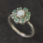 Opal Emerald Flower Ring worn on middle finger with light blue gradient nails. The opal shows flashes of fire colors surrounded by genuine emeralds in a floral cluster design. Perfect for adding inspiration and balance as an October birthstone and symbolizing love and growth as a May birthstone. Suitable for parties, birthdays, or casual outings.