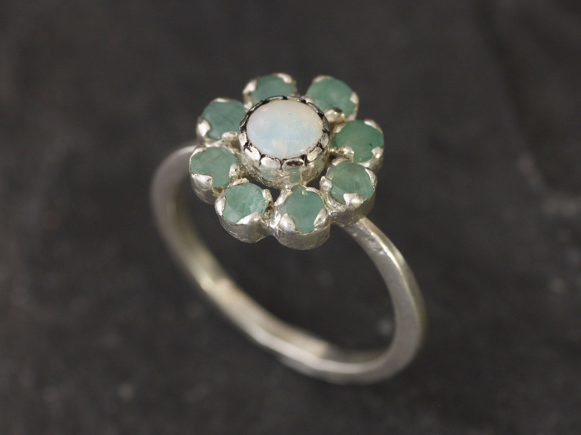Opal Emerald Flower Ring worn on middle finger with light blue gradient nails. The opal shows flashes of fire colors surrounded by genuine emeralds in a floral cluster design. Perfect for adding inspiration and balance as an October birthstone and symbolizing love and growth as a May birthstone. Suitable for parties, birthdays, or casual outings.