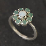 Opal Emerald Flower Ring worn on middle finger with light blue gradient nails. The opal shows flashes of fire colors surrounded by genuine emeralds in a floral cluster design. Perfect for adding inspiration and balance as an October birthstone and symbolizing love and growth as a May birthstone. Suitable for parties, birthdays, or casual outings.