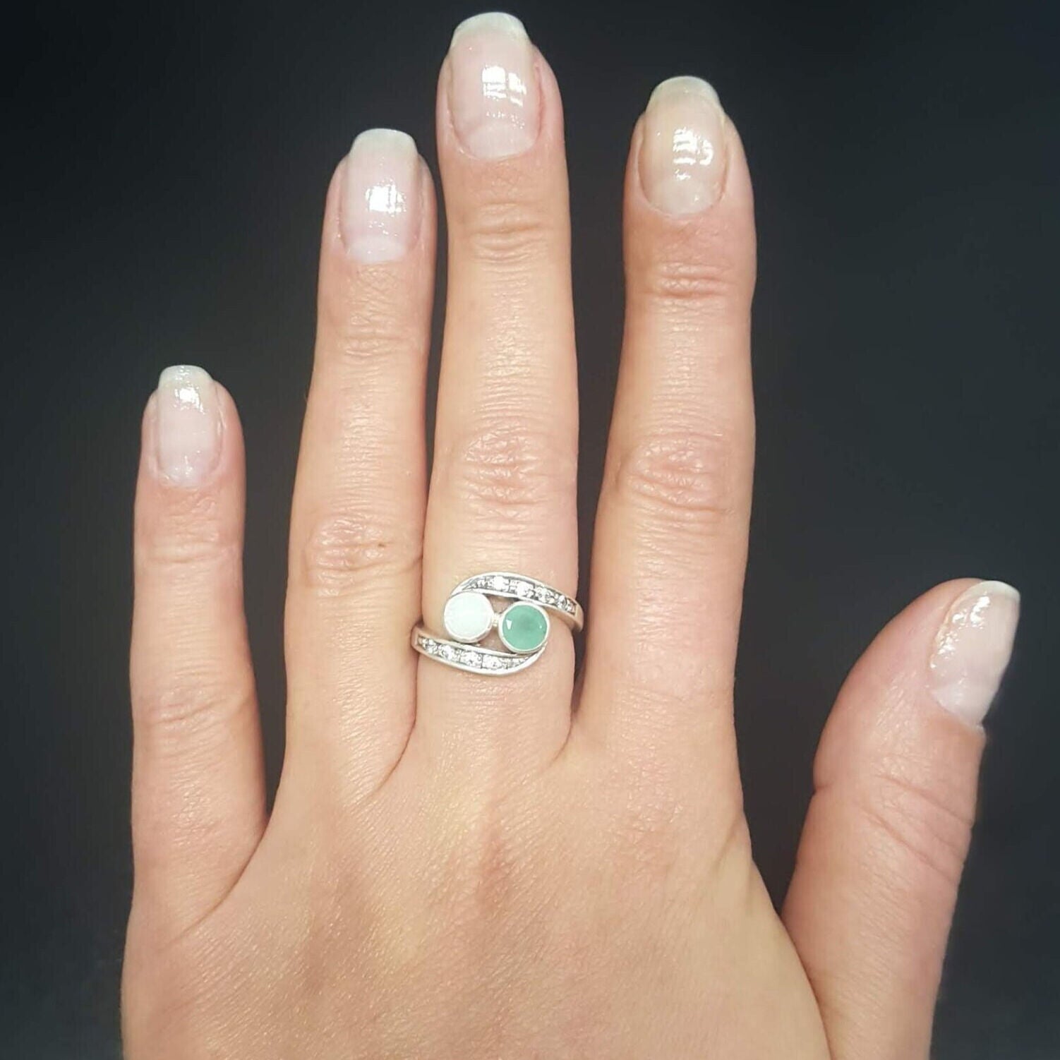 Bypass style ring featuring a natural Green Emerald and a cabochon White Opal in solid 925 sterling silver, accented with CZ pave stones for added brilliance. The combination of real Emerald and Opal highlights vibrant green tones and flashes of fire colors. With Emerald as the May Birthstone and Opal as the October Birthstone, this two stone design makes a meaningful gift for birthdays, anniversaries, and personal milestones. Perfect to wear at weddings, evening parties, and special celebrations.