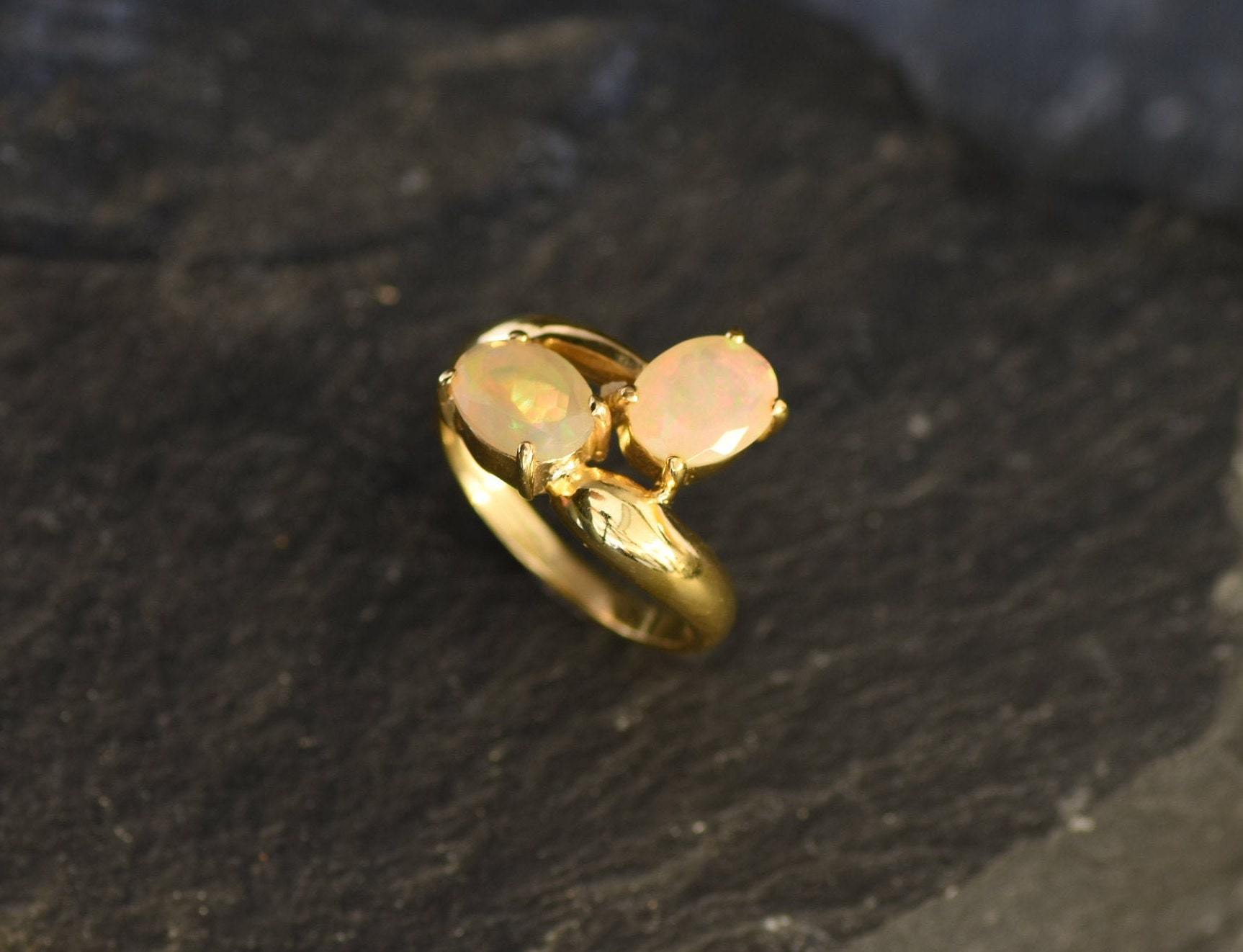 Two Stone Opal Ring in by pass style with twin cabochon gems showing flashes of fire colors, crafted as elegant fine jewelry. Known for promoting creativity and emotional healing, Opal is the October birthstone and a thoughtful gift for birthdays, anniversaries, and romantic occasions. Perfect to wear at weddings, evening events, and special celebrations.