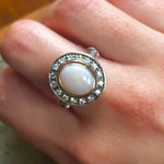 Opal Bezel Halo Ring worn on Middle  finger, showcasing an oval opal with rainbow and fire colors surrounded by CZ diamonds. Perfect for special occasions, weddings and cocktail parties. Ideal gift for October babies.