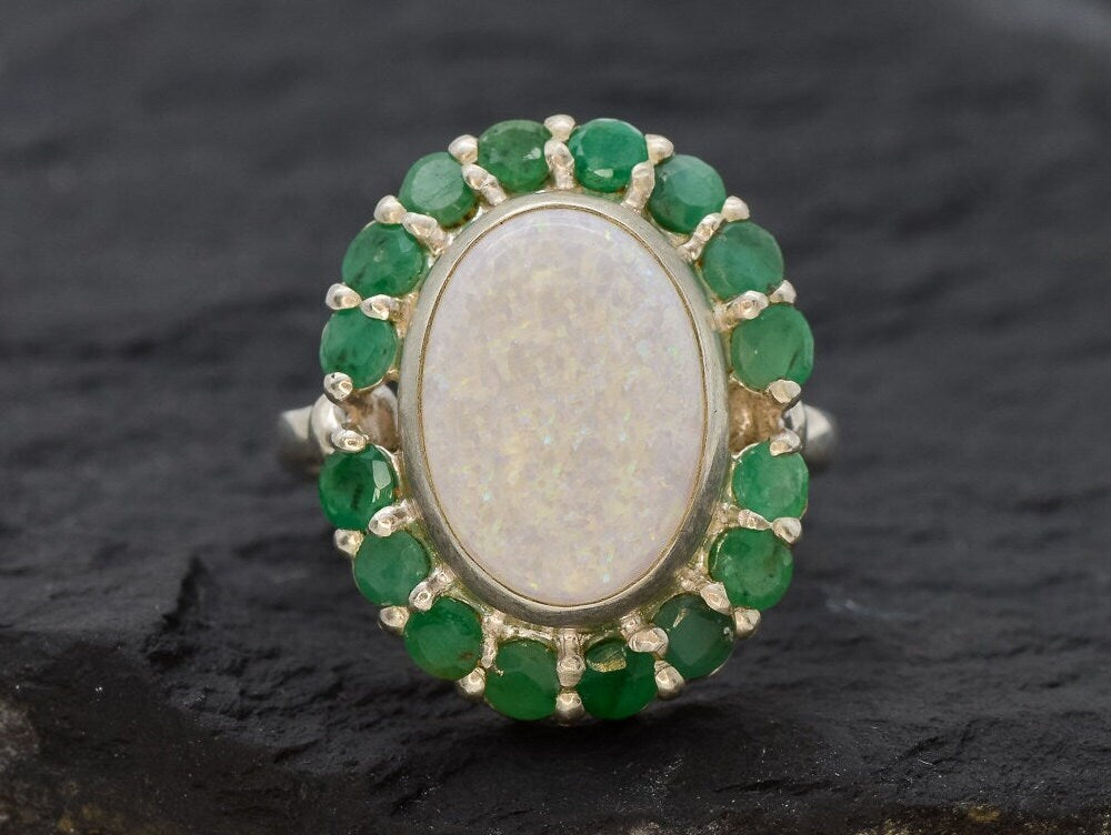 Opal and Emerald Large Cocktail Ring in sterling silver featuring an oval white opal cabochon framed with genuine emerald petals, a statement jewelry style that radiates flashes of fire colors while symbolizing love and renewal, making it a meaningful October and May birthstone gift idea. Perfect for weddings, cocktail parties, and formal evening events where a bold elegant look is desired.