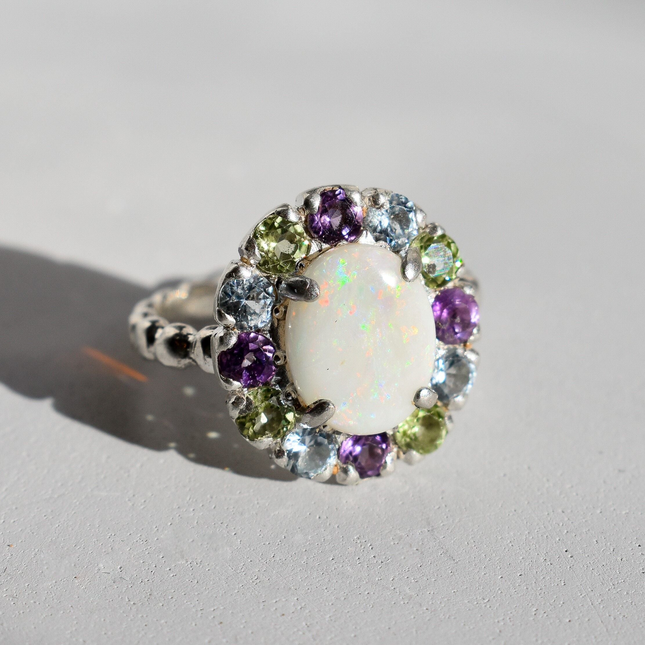 White Opal flower cluster ring in sterling silver with colorful Amethyst and Peridot halo in a cocktail style. The elegant cluster design adds sparkle. Opal is linked with hope and clarity, Amethyst with calm, Peridot with fresh energy. A lovely birthstone gift for October, February and August. Wear to weddings, graduations, dinner dates and holiday parties.