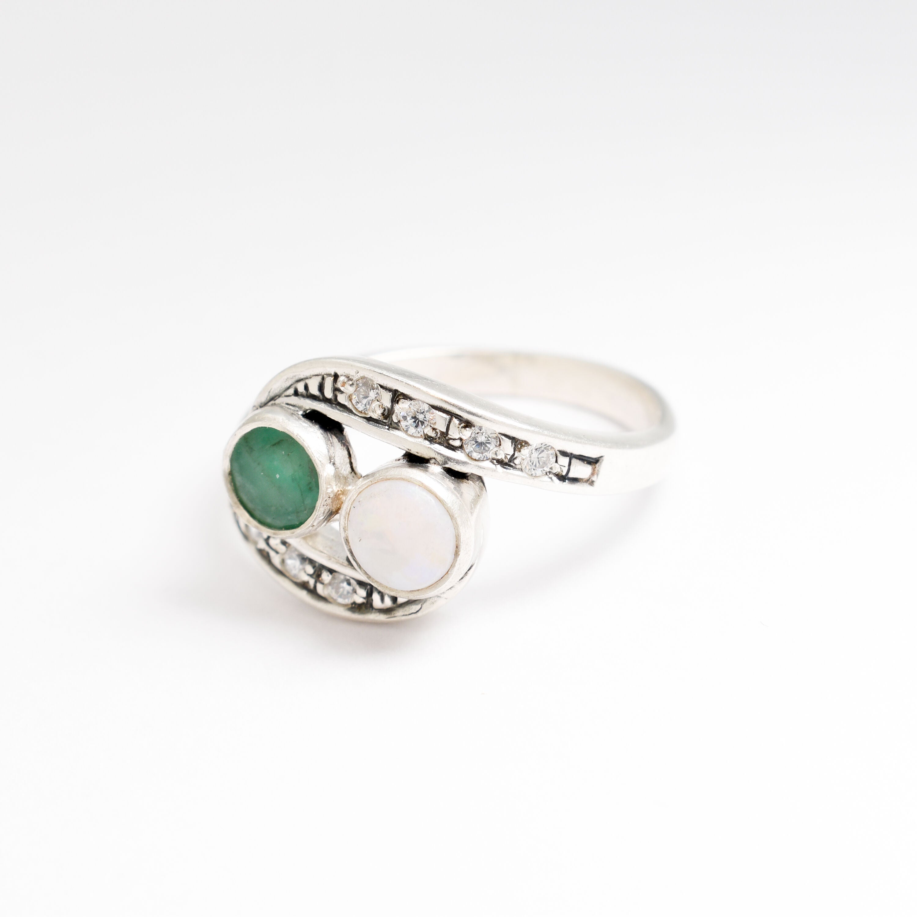 Bypass style ring featuring a natural Green Emerald and a cabochon White Opal in solid 925 sterling silver, accented with CZ pave stones for added brilliance. The combination of real Emerald and Opal highlights vibrant green tones and flashes of fire colors. With Emerald as the May Birthstone and Opal as the October Birthstone, this two stone design makes a meaningful gift for birthdays, anniversaries, and personal milestones. Perfect to wear at weddings, evening parties, and special celebrations.