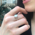 Opal Solitaire Ring styled with a black v-neck blouse. Jewelry style is elegant and modern with a bezel design showing flashes of rainbow colors. Symbolizes creativity, harmony, and positivity. A meaningful October birthstone gift for birthdays or milestones. Ideal for evening dinners, weddings, and cocktail events.
