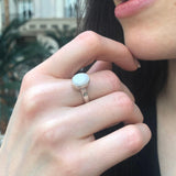 Opal Solitaire Ring styled with a black v-neck blouse. Jewelry style is elegant and modern with a bezel design showing flashes of rainbow colors. Symbolizes creativity, harmony, and positivity. A meaningful October birthstone gift for birthdays or milestones. Ideal for evening dinners, weddings, and cocktail events.
