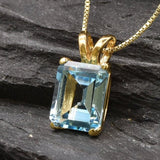 Emerald cut blue topaz pendant displayed on dark stone surface with reflective gold finish