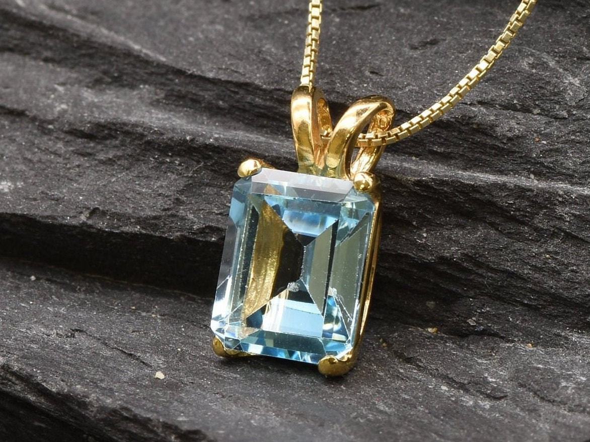 Emerald cut blue topaz pendant displayed on dark stone surface with reflective gold finish