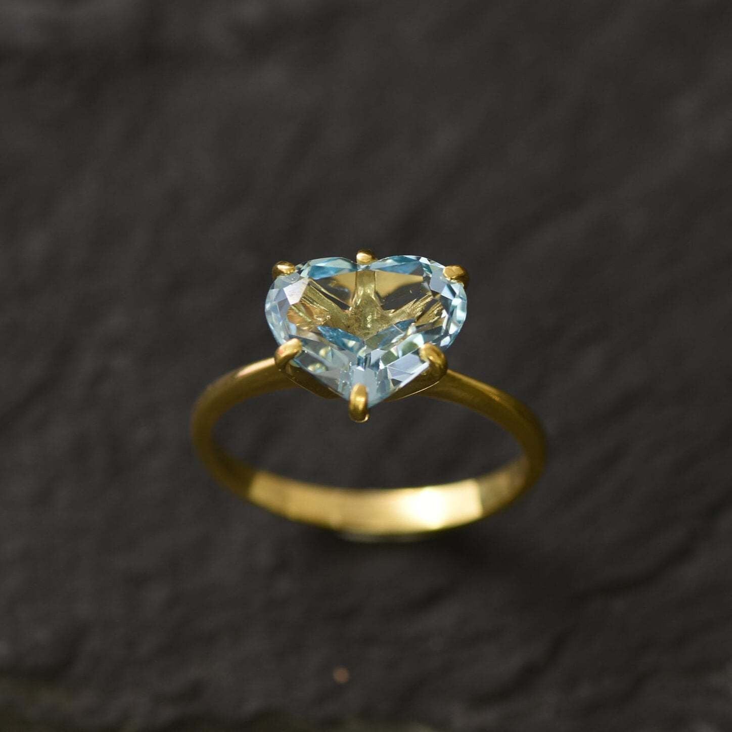 Side-angle close-up of blue topaz heart gold ring displaying gemstone details on dark surface