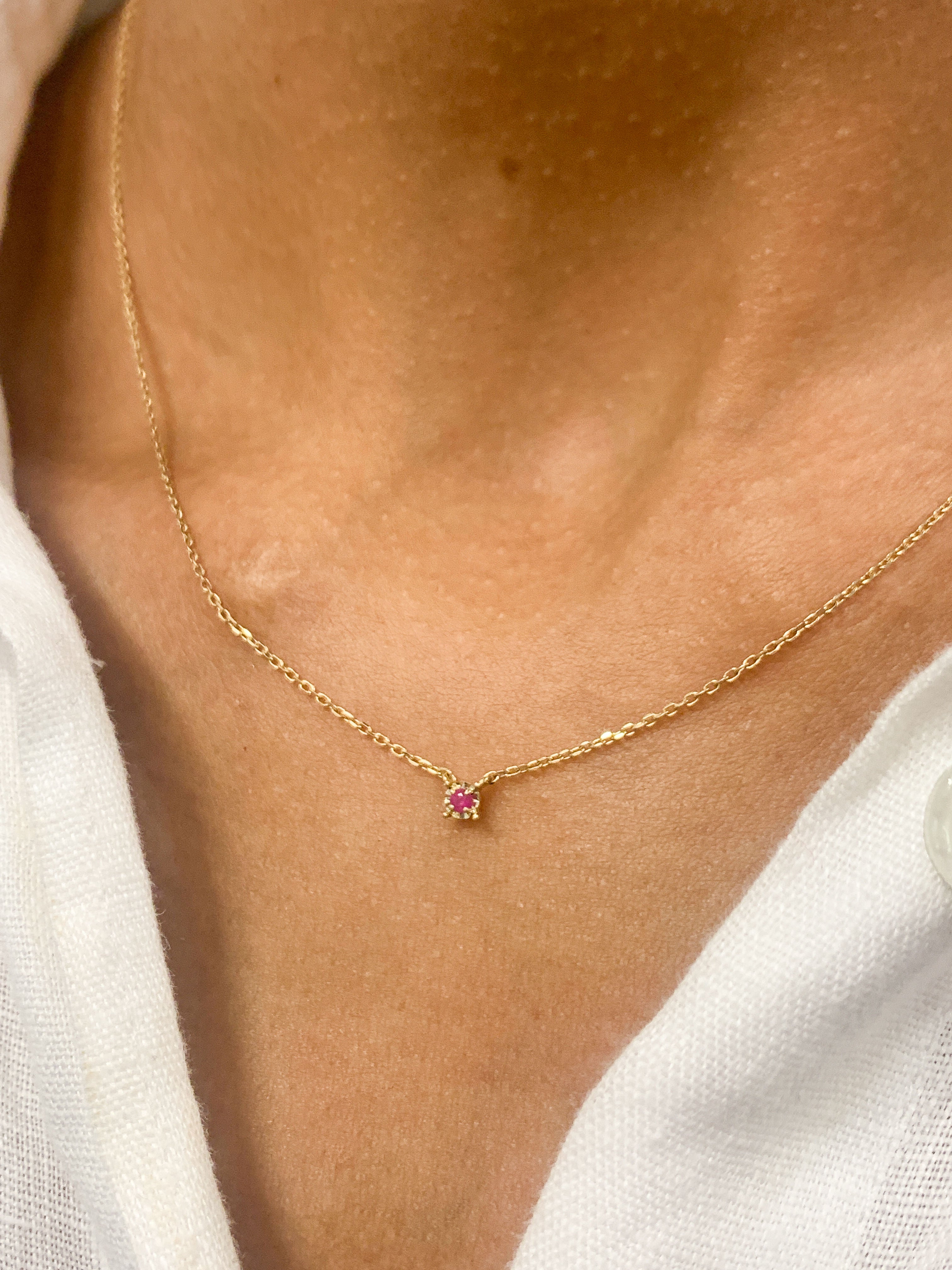 Woman in white top wearing a natural July birthstone Ruby necklace in solid 14k yellow gold chain. In a prong setting, and natural red color. Best worn in a minimalist look, can be worn as an everyday necklace