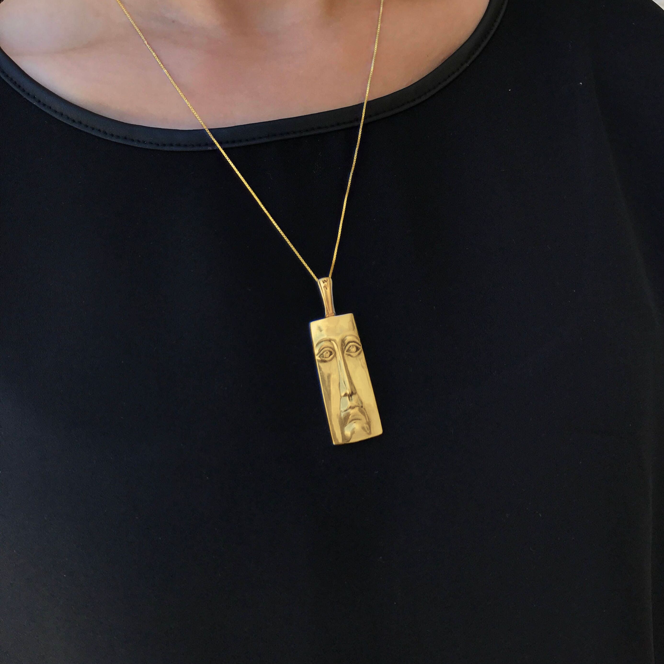 Woman wearing Long Face Pendant set in 18k yellow gold vermeil over solid 925 sterling silver. Gold statement pendant that matches well with black or darker outfit. Fits well for maximalist style.