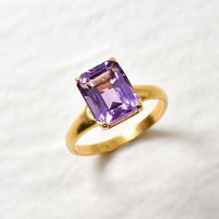 Yellow gold plated ring with amethyst February birthstone in emerald cut and prong setting. Bold solitaire ring ideal for special occasions and birthday gift. Encourages calmness, spiritual growth, and emotional balance. For peace, clarity, intuition